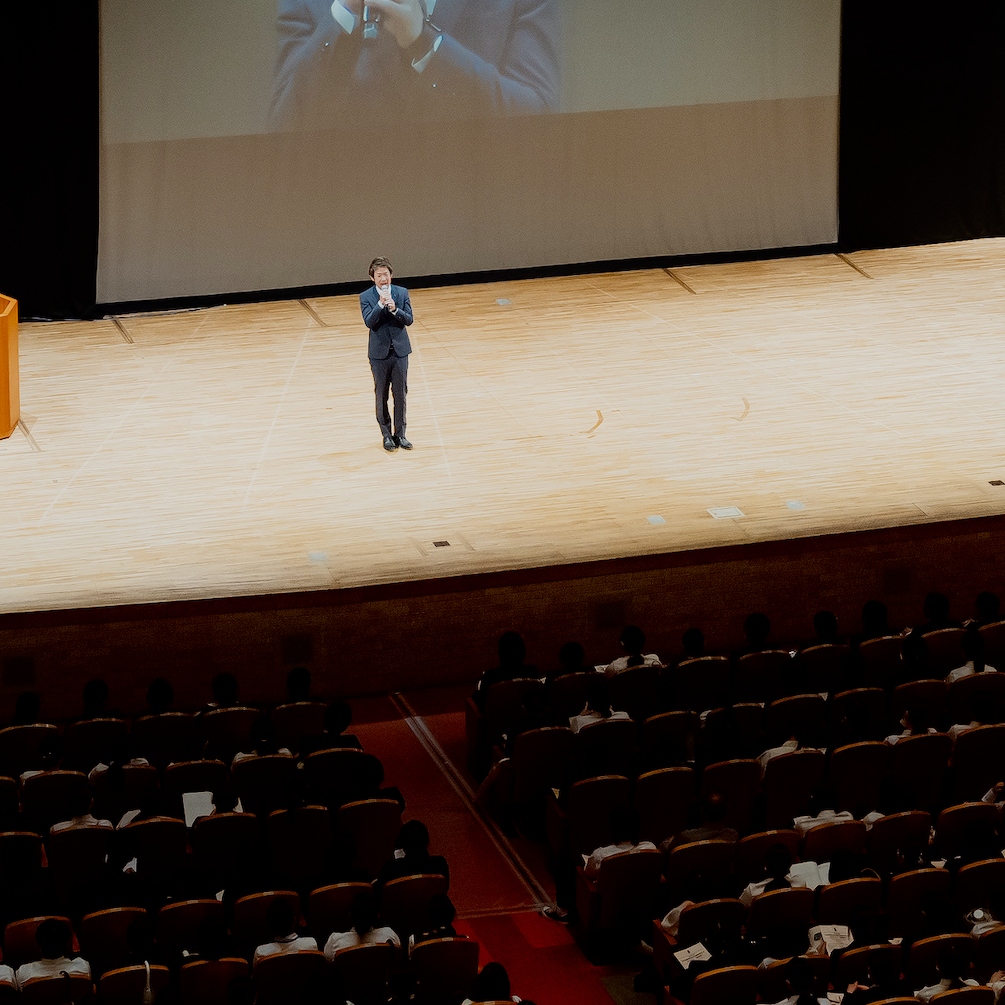 大きなホールのステージで講演する清水健と会場全景
