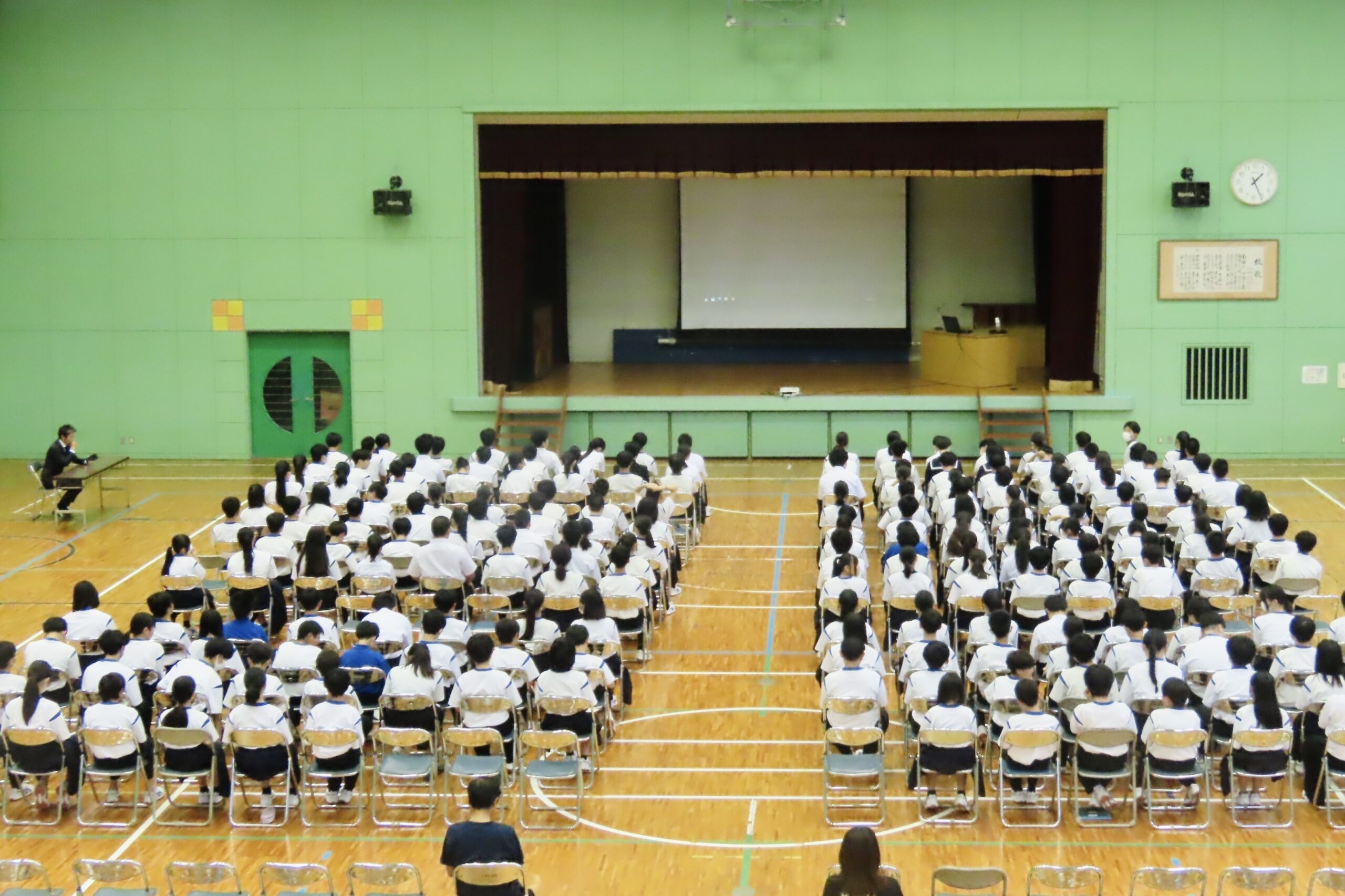 徳島県鳴門市立大麻中学校の体育館で開催された教育講演会。全校生徒が参加する会場の様子と、講演会の舞台。